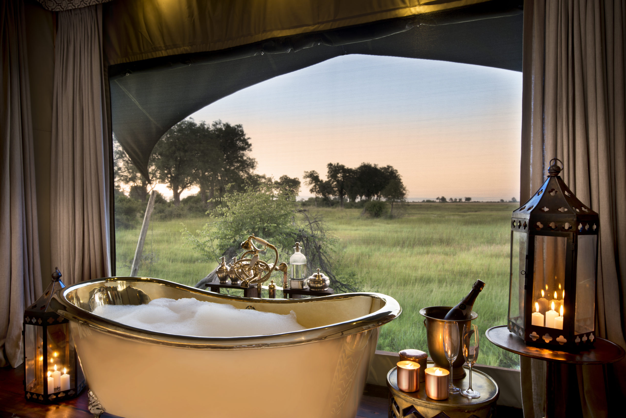 duba-plains-camp-rooms-luxury-tent-bathroom-02 A luxurious freestanding bathtub filled with bubbles sits beside candlelight, overlooking Botswana’s vast green plains—an indulgent way to experience the best time to visit Botswana for a safari.